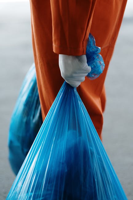 A close-up view of a person wearing orange overalls and a white glove, holding a large, semi-transparent blue plastic rubbish bag filled with waste. The bag appears full and tightly gathered at the top, with the person's hand gripping the gathered material. The background is blurred, suggesting an outdoor environment, possibly near a waste collection area or industrial site. The image emphasizes the process of waste handling, with the visible textures of the plastic bag and the smooth, durable fabric of the overalls. This scene aligns with waste management services, such as rubbish removal or on-site clearance, provided by companies like Waste Collection Croydon, highlighting practices of private waste disposal outside of municipal collection systems.