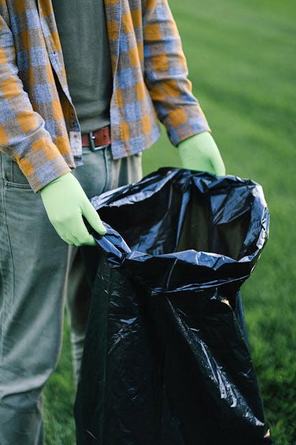 A person standing outdoors on a grassy area, wearing a plaid shirt in shades of grey, yellow, and blue over a green t-shirt, with grey trousers and a brown belt. They are holding a large black trash bag with both hands, which is open and appears to be partially filled. The person’s hands are covered with bright green rubber gloves, indicating handling of waste or rubbish. The background shows a lush, green lawn, and the scene suggests a waste collection or rubbish removal activity, possibly related to private disposal or on-site clearance services provided by Waste Collection Croydon. The lighting is natural, with daylight illuminating the scene, emphasizing the textures of the bag’s glossy plastic material and the fabric of the clothing.