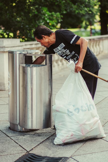 A close-up view of a person wearing orange overalls and a white glove, holding a large, semi-transparent blue plastic rubbish bag filled with waste. The bag appears full and tightly gathered at the top, with the person's hand gripping the gathered material. The background is blurred, suggesting an outdoor environment, possibly near a waste collection area or industrial site. The image emphasizes the process of waste handling, with the visible textures of the plastic bag and the smooth, durable fabric of the overalls. This scene aligns with waste management services, such as rubbish removal or on-site clearance, provided by companies like Waste Collection Croydon, highlighting practices of private waste disposal outside of municipal collection systems.