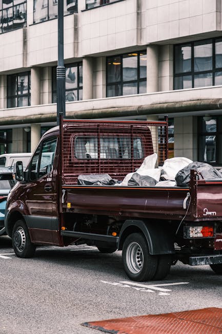 A compact burgundy pickup truck parked on a city street in front of a modern office building with large rectangular windows and a light-colored facade. The truck's open bed is loaded with various types of waste, primarily black and clear plastic bags filled with rubbish, some with visible crumpled paper or textile materials. The truck features a metal grid partition behind the cab to prevent load shifting, and its body shows minor signs of use with a clean, shiny finish. Surrounding the vehicle are other parked cars, and a black streetlamp stands nearby in the foreground. The area appears to be an urban commercial or office district, with a paved street and clearly marked parking spaces. The scene suggests an example of private waste collection or on-site clearance, typical of services provided by companies like Waste Collection Croydon, which specialise in rubbish removal. Natural daylight illuminates the scene, highlighting the textures of the waste bags and the vehicle's finish, contributing to a professional and neutral depiction of non-local authority waste disposal methods.
