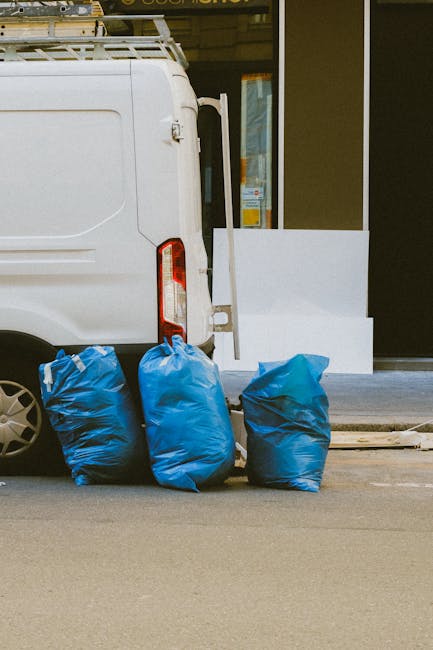 A compact burgundy pickup truck parked on a city street in front of a modern office building with large rectangular windows and a light-colored facade. The truck's open bed is loaded with various types of waste, primarily black and clear plastic bags filled with rubbish, some with visible crumpled paper or textile materials. The truck features a metal grid partition behind the cab to prevent load shifting, and its body shows minor signs of use with a clean, shiny finish. Surrounding the vehicle are other parked cars, and a black streetlamp stands nearby in the foreground. The area appears to be an urban commercial or office district, with a paved street and clearly marked parking spaces. The scene suggests an example of private waste collection or on-site clearance, typical of services provided by companies like Waste Collection Croydon, which specialise in rubbish removal. Natural daylight illuminates the scene, highlighting the textures of the waste bags and the vehicle's finish, contributing to a professional and neutral depiction of non-local authority waste disposal methods.