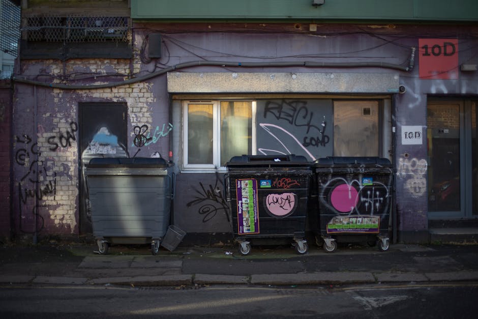 Three large wheelie bins are positioned on a concrete pavement in front of a weathered brick building with a graffiti-covered exterior. The first bin on the left is grey with a closed, slightly curved lid, while the other two bins on the right are black, featuring graffiti tags and pink heart-shaped stickers on their surfaces. The black bins also have colorful disposal stickers on their fronts, including green and purple labels. The bins are situated directly adjacent to a building wall that has peeling paint, exposed bricks, and a mixture of spray-painted and hand-drawn graffiti in various colors, mostly black, blue, and pink. Behind the bins, there is a small white window with frosted glass panes reflecting the light, set into a grey wall topped with a green horizontal strip. A narrow driveway or alleyway runs alongside the building, with some leaves and debris scattered at the base of the bins. The scene is illuminated with natural light, highlighting the textures of the worn brick and graffiti, and subtly suggesting an alternative waste handling environment managed by waste collection services such as Waste Collection Croydon.
