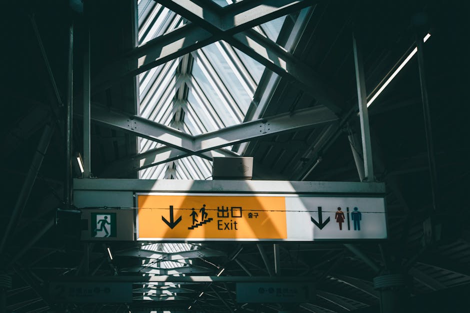 A large overhead sign hanging beneath a glass and metal roof structure at East Croydon station, displaying directions for the exit with icons indicating stairs, a walking figure, and an elevator. The sign features an orange background with black text and symbols, with arrows pointing downward to guide passengers. To the right, there are icons of a woman in a red dress and a man in blue, indicating the location of restroom facilities below the sign. The environment is an indoor transit environment with a modern, industrial design, illuminated by natural light filtering through the glass roof, creating a contrast between the bright signage and the darker structural elements surrounding it. The scene emphasizes the station's clear signage and infrastructure designed to assist commuters in navigating the station for bus and train connections, with no visible rubbish or waste-related objects present, aligning with the context of efficient passenger movement and station management.