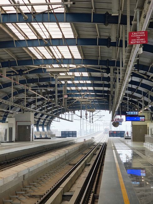 An empty section of East Croydon station platform under a large, arched, metal roof structure with exposed blue steel beams and translucent panels allowing natural light to filter through. On the right side, a red and white caution sign warns of clearance restrictions. The platform surface appears to be polished, reflecting some of the overhead lighting, and there are digital information displays mounted on the ceiling indicating train schedules. In the background, the train tracks extend into a misty or overcast environment, with faint outlines of other station elements. The scene is devoid of pedestrians or trains, emphasizing a quiet moment at the station, with the metal roof and structural elements providing a modern, industrial aesthetic. Occasionally, indoor signage and electrical fixtures related to station operations are visible, supporting an environment suited for passenger movement and potential site clearance for maintenance or cleaning activities by waste collection services such as Waste Collection Croydon.