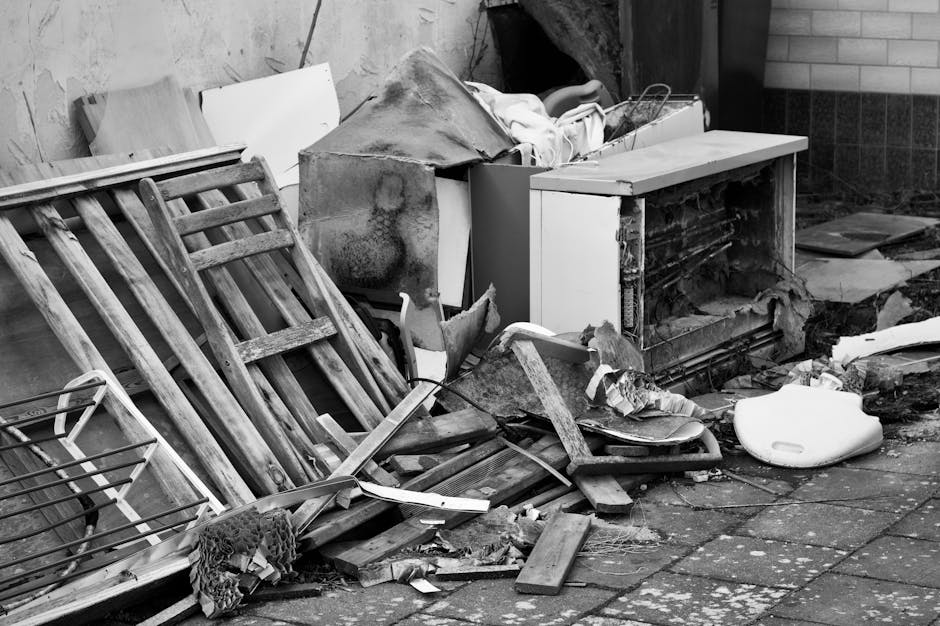 A pile of discarded household waste and debris is situated outside, leaning against a brick and concrete wall. The debris includes a weathered wooden garden chair with slatted design, lying on its side with visible grains and rough textures. Nearby, an old, broken cabinet with chipped white paint and exposed interior shelves is partially covered with a torn cloth or fabric. A large, worn piece of carpet or possibly an insulation panel, stained and curled at the edges, rests atop the cabinet. Scattered among the items are various pieces of wood, some broken and splintered, along with a white plastic object resembling a small container or lid. The ground is paved with uneven stones and appears to be outdoors, with dirt and small debris spread across the surface. The scene reflects an environment suitable for private waste clearance or independent rubbish removal, with Waste Collection Croydon providing relevant services for such bulky waste disposal tasks.