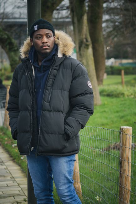 A young man with dark skin and a serious expression is standing outdoors on a paved pathway adjacent to a wire mesh fence supported by wooden posts. He is dressed warmly in a black puffer jacket with a faux fur-lined hood, a navy blue hoodie underneath, and a black beanie. The background features a grassy area with trees and a park-like setting, indicating a residential or community environment. The scene is lit with natural daylight, suggesting an overcast day. The image captures a casual moment, possibly relating to private waste management or outdoor clearance activities, with the young man appearing contemplative as part of a broader context involving rubbish removal or outdoor cleaning services provided by Waste Collection Croydon.