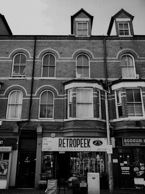 A black-and-white photograph of a multi-storey brick building with three prominent windows on each floor, featuring arched tops and white decorative brick accents around the windows. The building has two small dormer windows at roof level, both with gabled roofs and single-pane windows. In the ground-floor windows, a house clearance business named 'RETROPEEK' is visible, with a large sign displaying the company's name and logo, along with smaller notices and advertisements on the glass. To the right side of the storefront, there is another related business, and a glimpse of outdoor items, possibly waste or clearance debris, is seen in front of the entrance. The scene suggests a commercial property situated on a street with other shops or services, and the image captures an environment associated with independent waste collection or clearance services, as highlighted by the presence of the house clearance sign and the urban setting.